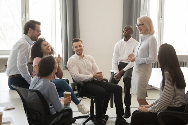 A diverse group of employees sit casually in an office, smiling and talking with their manager.