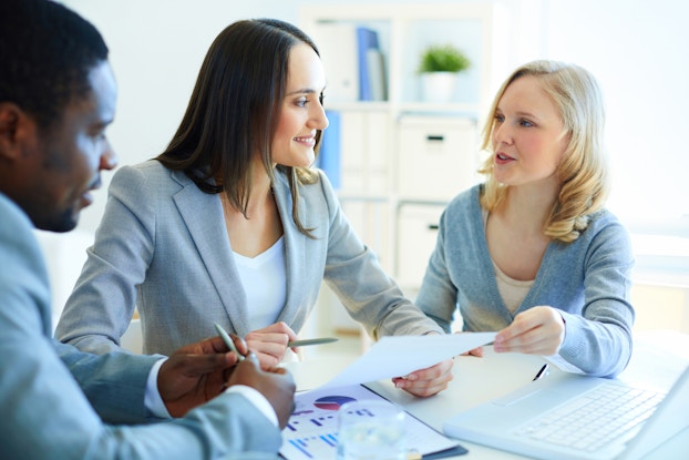 Three people sit around a table in a brightly lit room with white walls. The man on the left is partially out of frame and out of focus. On the table between him and the woman in the middle is a clipboard with a paper showing several different graphs in various colors. The woman in the middle wears a light gray blazer and has dark hair, while the woman on the right has blonde hair and wears a light blue cardigan. The two women are looking at each other and the woman on the right is speaking as she hands a piece of paper to the woman in the middle.