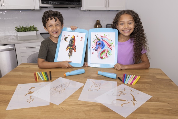 A young boy and girl sit at a kitchen table holding up stenciled artwork created with Flo-Magic's stencil and airbrush kits. The boy on the left holds up artwork of a rocketship and the girl on the right holds up artwork of a unicorn. The Flo-Magic stencils, airbrushes, and markers are arranged on the table in front of the kids.