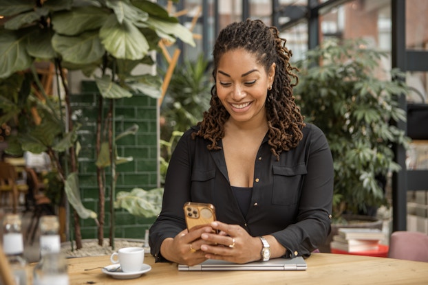 A woman sits at a table in a cafe or restaurant and looks down at the smartphone in her hand with a smile. She has dark braided hair that reaches her shoulders and she wears a black shirt with gold buttons. Behind her are several large, leafy potted plants.