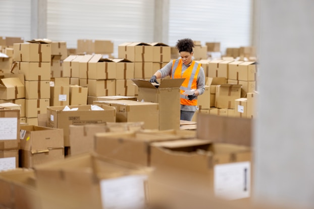 A warehouse employee stands in a sea of cardboard boxes, some of them open. The employee, who wears a bright orange high-visibility vest and rubber gloves, is opening one of the boxes while glancing at a piece of paper in her hand.