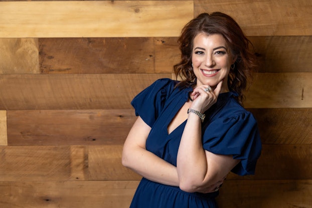 Amber Gaige, Founder of Far Beyond Marketing, smiles as she poses with her arms folded and one hand under her chin. She is a white woman with wavy brown hair and brown eyes. She wears a dark blue blouse with loose butterfly sleeves.