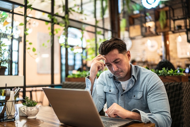 A young man in a chambray button-up shirt sits at an open laptop at a table. The man holds a loose fist to his forehead as he looks at the laptop screen with a worried look on his face. On the table next to his laptop is a succulent plant in a small white vase and a black wire holder filled with utensils and folded napkins. The room around the man is a large glass-walled atrium with vines dangling from the ceiling