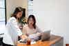 Two Asian women business partners are pictured in a well-lit modern office. One of them is holding a financial report and the two are discussing a financial matter relating to their company.