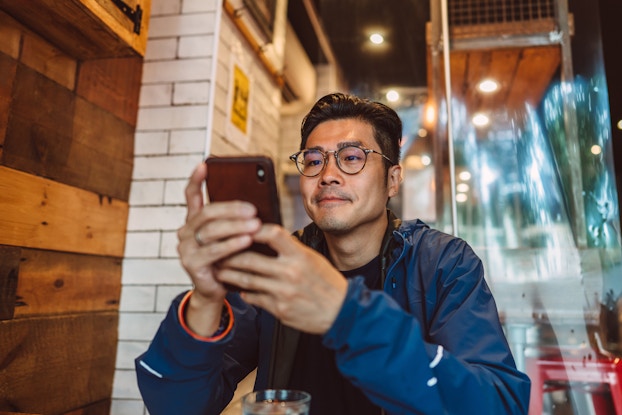 A young man sits with his elbows on the table and looks at the smartphone in his hands. The man wears a dark blue windbreaker and glasses. He has neatly combed black hair and is looking at his phone with a neutral expression.