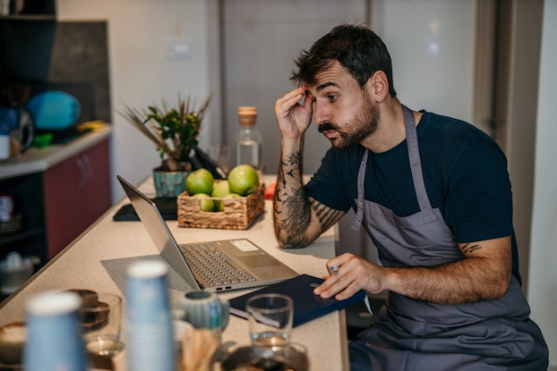 A concerned restaurant owner is seated at the restaurant bar looking at a laptop computer placed on the counter. He is reviewing his restaurant's finances.