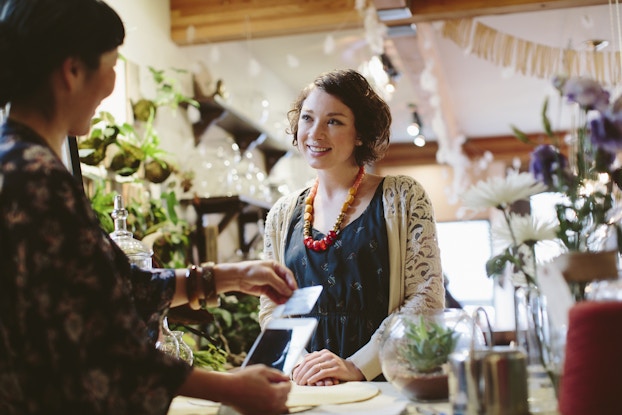 A young woman with bobbed, wavy auburn hair stands at the checkout desk in a shop filled with potted plants. She wears a dark blue dress, cream cardigan, and a necklace made of chunky orange beads. Her hands are folded on the desk and she smiles at the cashier, who faces away from the viewer and holds a credit card above a digital tablet to complete the transaction.