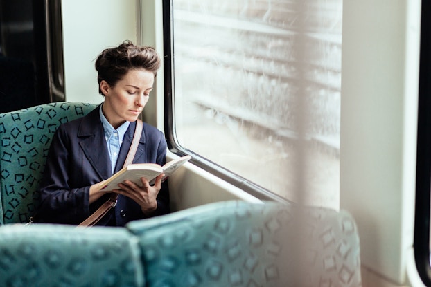 A young woman is seated on a train by a window. She is holding a book in her hands and is reading.