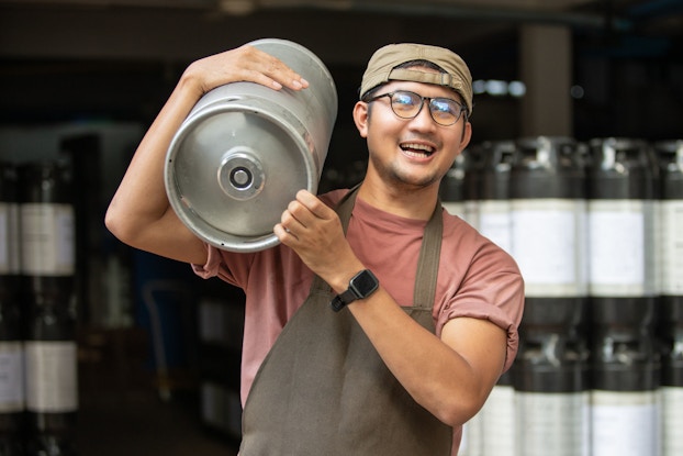 A male brewer is pictured smiling and carrying a keg of beer.
