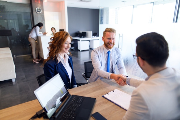 A couple sits across a desk from a man in glasses. The members of the couple, a man and a woman, are both wearing businesswear and smiling; the man reaches across the desk and shakes the hand of the man in glasses. On the desk are an open laptop and a clipboard. In the background is an open plan office; a woman leans on a desk near the back wall and looks at her smartphone while another woman sits behind the desk.
