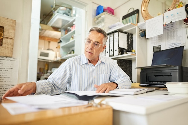 An older man is seated at a desk in his office. He has papers spread out in front of him. He is reviewing a list of expenses he can claim as deductions for his business taxes.