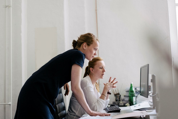 Two women are pictured in an office setting. They are both leaning over a desk looking at a computer screen. One of the women is illustrating a point by gesturing with her hands.