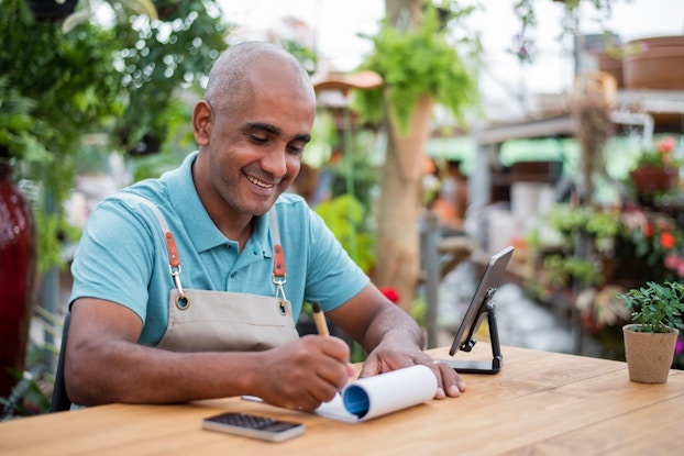 A bald man wearing a teal collared shirt under a tan apron sits at a wooden table outside. He smiles as he writes something on a notepad. Beside the notepad on the table are a smartphone on a stand and a small calculator. Behind the man are lines of potted plants and a cluster of trees, with an out-of-focus building standing a greater distance away.