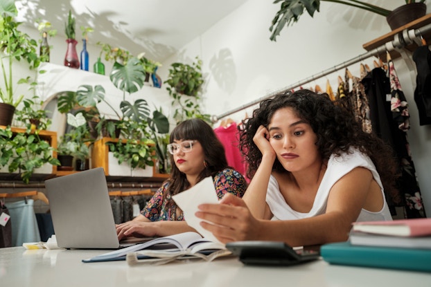 Two young women co-business owners of a clothing store sit at table and do their business's bookkeeping. Behind them is a rack of merchandise.