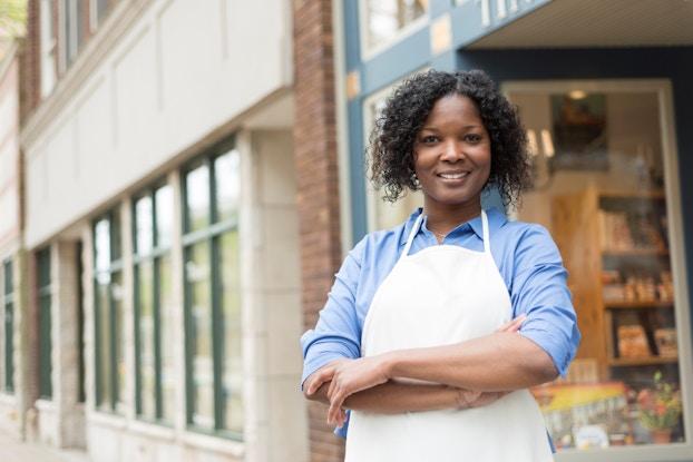 A woman small business owner stands outside of her shop. She is smiling at the viewer with her arms folded in front of her. Her shop is located on a main street in a U.S. small city.