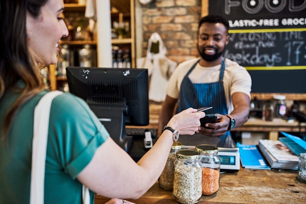 A woman uses tap on a credit card reader to pay for groceries at a neighborhood market.