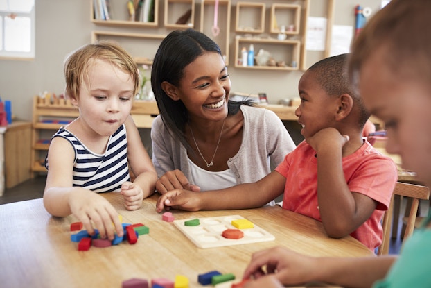 A woman sits between two children of about 3 or 4 years of age around a low wooden table. The child on the left is grabbing at one of the 3D shapes in the pile in front of him. The child on the right is grinning at the adult woman, who smiles widely back at him. This child has a puzzle in front of him with nine spaces for nine different shapes. Three of the spaces are filled with shapes (octagon, semicircle, and square) from the pile on the table. In the foreground, a third child looks down at his own pile of shapes.