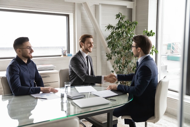 Three men sit around a glass-topped conference table in a white-walled office room. The two men on the right are wearing suits; they sit across from each other and shake hands. The man on the left is wearing a dark blue button-up shirt; he watches them with a smile.