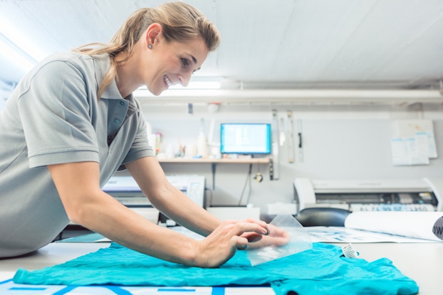 A smiling woman peels a clear sheet of plastic off of the front of a teal T-shirt as part of the shirt printing process. A design in white can be seen underneath the plastic sheet.