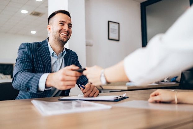 A businessman is seated at a desk. He is smiling as he prepares to sign loan documents for a small business loan.