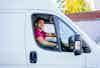 A man is seated in the driver's seat of a delivery van. He is wearing a hat and smiling at the viewer. He is in the process of making deliveries for his employer.