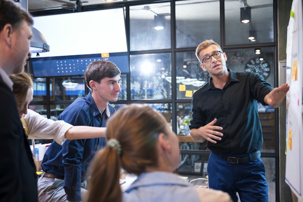 On the right side of the photo, a man in glasses and a black button-up shirt stands next to a white board covered with post-it notes. The man is speaking and gesturing to something on the board with one hand. To the left of him, several other people watch him and look at the board. In the background is a floor-to-ceiling glass wall, on which is mounted a flat screen TV. On the other side of the wall is an open warehouse or office space with cylindrical lights hanging from the ceiling.
