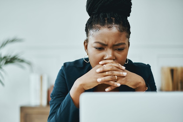 A young woman is staring at her laptop computer with a concerned look on her face. Her brow is furrowed and she is holding her hands in front of her mouth while she tries to troubleshoot a computer problem.