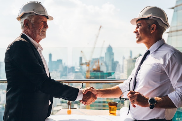 Two men in white hard hats shake hands. Behind them is a city skyline with a bright yellow crane working on one of the buildings.