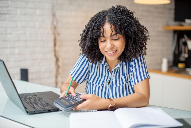A young woman is seated at a desk in a modern office space. She has a laptop open and a hard copy of an accounting ledger in front of her. She is holding a calculator in her hands and updating her accounting records.