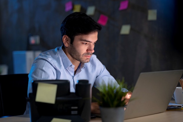 A young business owner sits in a darkened office at night. He is looking at his laptop computer and analyzing data.