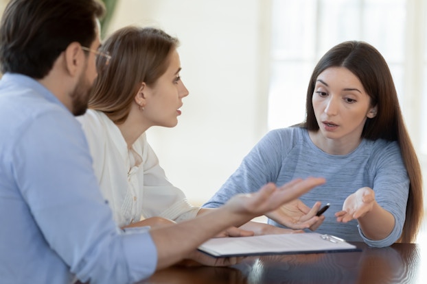 A young woman has a tense conversation with a couple sitting at a table with her. The young woman is seated on the right; she has long dark hair and holds a pen in one hand. She gestures with both hands at a clipboard on the table. The couple speaking with her are a man and a woman seen in profile. The man has one hand up in a gesture of confusion.