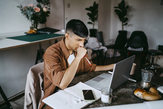 A young man sits at a table, looking at the screen of his laptop with one fist held to his mouth in worry. The man has dark hair with shaved sides and he's wearing a brown shirt over a white T-shirt. An open notebook and a smartphone sit next to the laptop.