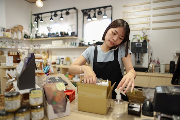 A woman employee of a eco-friendly home goods store is pictured preparing an order for shipment. She is holding her phone to her ear while packing the box with items to be shipped.