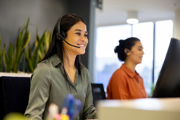 A young customer service representative is smiling and wearing a headset with a microphone. She is answering a customer's question over the telephone.