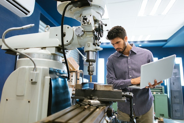 A man holding a laptop stands next to a table-top drilling machine and watches it drill a hole in a hollow block of metal. The machine is large and white, with a short arm extending over a metal platform. The platform has grooves in it that allow objects to be screwed in and held still while they are drilled. Various cubes and squares of metal are currently screwed into the platform. The man next to the machine has dark hair and a dark beard, and he is wearing khakis and a purple-and-white windowpane-patterned button-up shirt.