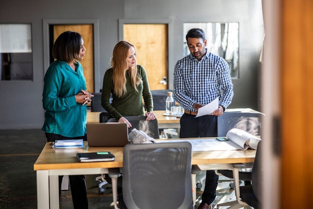 A group of work colleagues stand before a desk and discuss an issue. Behind them is a modern office setting.