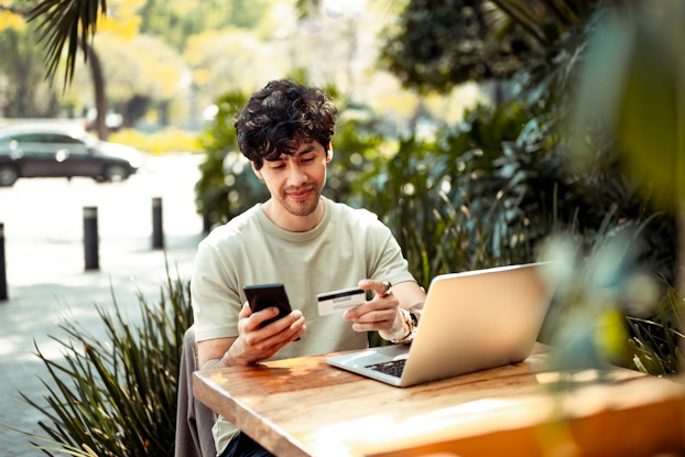 A young man sits at a wooden table outside, surrounded by plants with a parking lot in the background. He holds a credit card in one hand and a smartphone in the other, and a laptop sits open on the table in front of him.