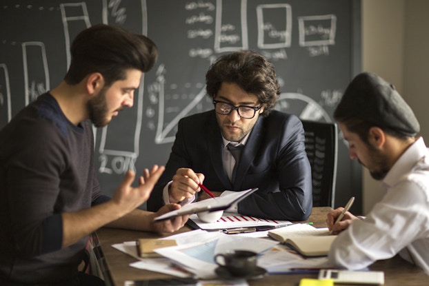 Three men sit around a table piled with papers. The man on the left, who has dark hair and a dark beard, holds out a clipboard. The man in the middle, who has dark hair and glasses, points at something on the clipboard with a red pencil. The man on the right wears a black beret on the back of his head; he writes something in a notebook. In the background is a blackboard with several graphs drawn on it in chalk.
