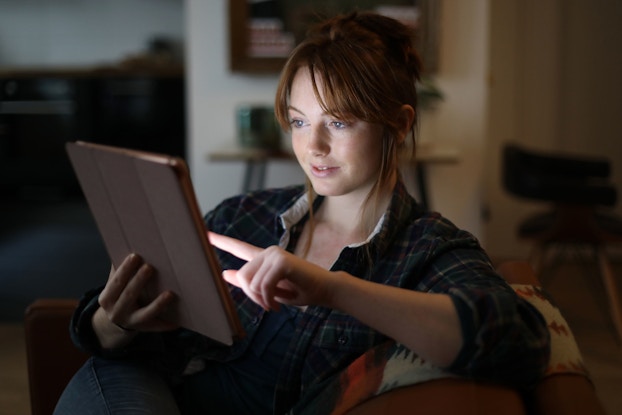 A young woman is sitting in her home. She is holding a tablet and perusing online goods. Her face is illuminated by the glow of the tablet.