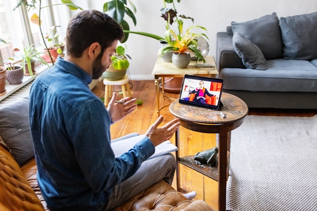 A man sits in a leather armchair facing away from the viewer and toward an electronic tablet propped up on top of a small wooden end table. On the screen of the tablet is the image of a woman sitting in an orange armchair; she has one hand to her chin and appears to be listening thoughtfully. The man has his hands up in a gesture, as if he is speaking to the woman on screen. Both the man and the woman have a packet of papers on their laps.