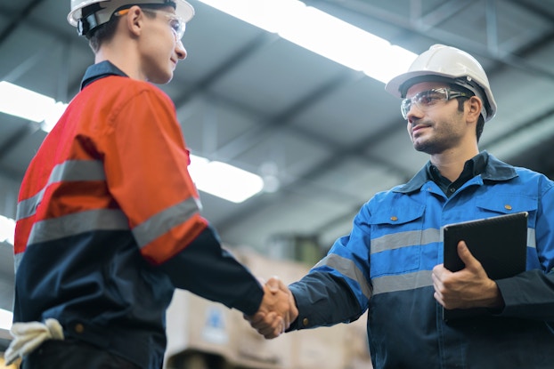 An angled picture shot from below, showing two young men shaking hands. Both men wear safety glasses and white hard hats. The man on the left wears a red-orange hi-visibility windbreaker; the man on the right holds a clipboard and wears a bright blue version of the windbreaker. The men are standing in a warehouse; the photo shows the flourescent light strips on the ceiling.