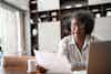 A middle-aged woman wearing glasses and a black-and-white blouse sits at a wooden table and considers the piece of paper that she holds in her hand. In the background is a living room or den, with dark wood shelving along the back wall. An open laptop sits in the foreground, in front of the woman.