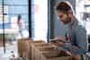 A man has several boxes lined up resting on top of a title. He is preparing several orders for shipment. He holds a computer tablet in his left hand to double-check orders as he prepares them.