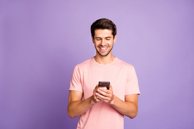 Person standing in front of a neon purple background smiling down at a phone.
