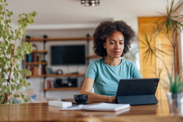 Woman works on laptop in home office with coffee mug and notebook on her desk.