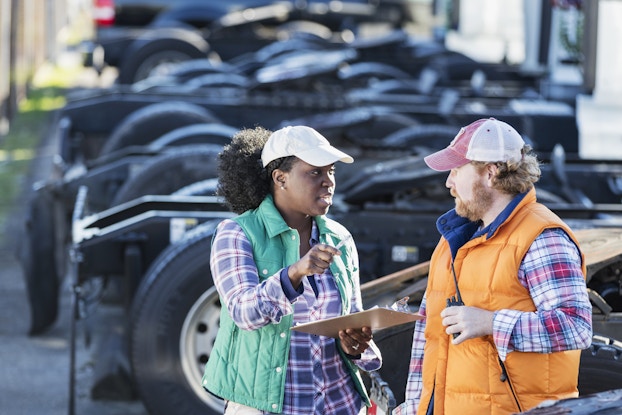 Two fleet management employees have a conversation in front of a line of semi-trucks. The employee on the left is a woman with dark curly hair in a ponytail. She wears a blue plaid shirt under a green puffy vest and a white baseball cap, and she holds a clipboard in one hand while gesturing with a pen with the other hand. The employee on the right is a man with a reddish-brown beard. He wears a red-and-blue plaid shirt under an orange puffy vest and a red-and-white baseball cap.