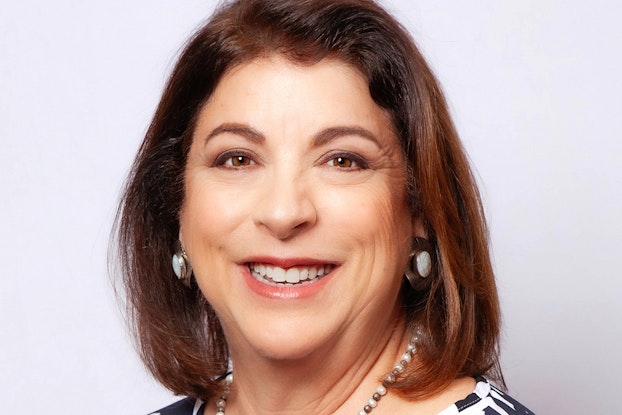 A head shot of Fran Maier, Founder of Match and Founder and CEO of BabyQuip. Fran is a white woman with shoulder-length brown hair and brown eyes. She is smiling and wearing a black-and-white patterned shirt, a gray-and-white pearl necklace, and dangling gold earrings with white stones.