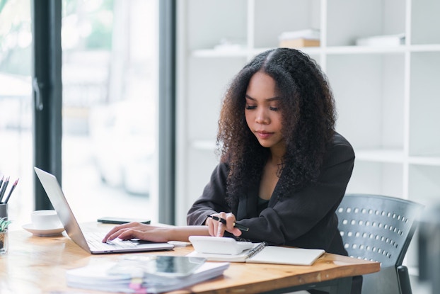 A young woman is seated at a desk in her modern office. She has her laptop in front of her, a calculator, and an open notebook. She is entering calculations into the accounting program to keep her business's books updated.