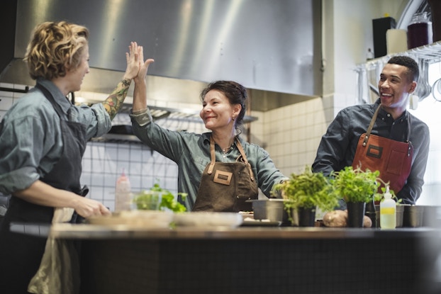 A trio of restaurant employees, each wearing an apron over a blue-gray collared shirt, stand behind a prep table in a kitchen with tiled walls. The two people on the left high-five while the man on the right looks on. The prep table is covered with empty plates, condiment bottles, and pots of greens.