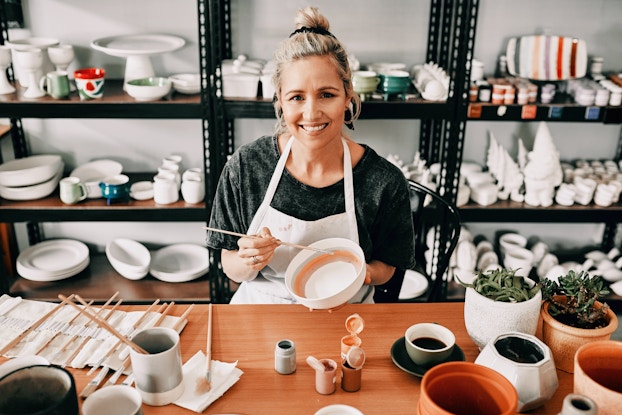 A young woman sits at a long wooden table in front of a set of black metal shelves. Both the shelves and the table are filled with white pottery in a variety of shapes. The table also holds a row of paintbrushes in a roll-up cloth holder. The woman is wearing an apron and holding a bowl and a long, thin paintbrush. The bowl has a few lines of blue, orange, and mauve painted around its inside.